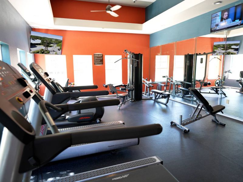 Elm Hollow apartments Modern gym with treadmills, weight machines, mirrors, and TVs on orange and blue walls, brightly lit by windows.