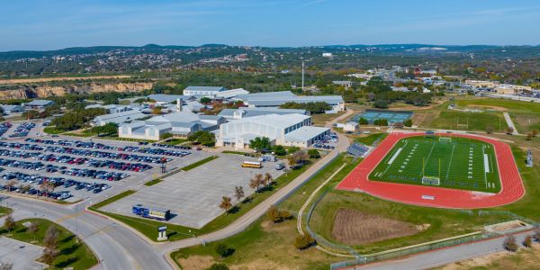 Aerial view of a large campus, featuring Elm Hollow Apartments alongside buildings, a parking lot, and a sports field with a track under the clear blue sky.