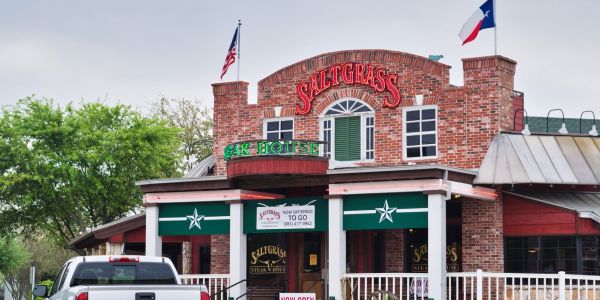 Brick steakhouse with a "Saltgrass" sign, U.S. and Texas flags, green awnings, and a parked white truck, nestled just around the corner from Elm Hollow Apartments.