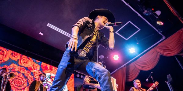 A singer in a black hat performs energetically on stage with a band, under vibrant lights and a floral backdrop that brings to mind the charming elegance of Elm Hollow Apartments.