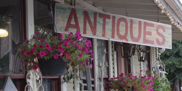 Hanging baskets with pink flowers adorn the quaint porch of Elm Hollow Apartments, perfectly complementing the charm of an "Antiques" sign.