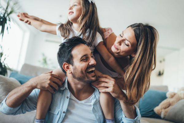 A joyful family moment unfolds at Elm Hollow Apartments, with a father carrying a child on his shoulders and the mother smiling beside them indoors.