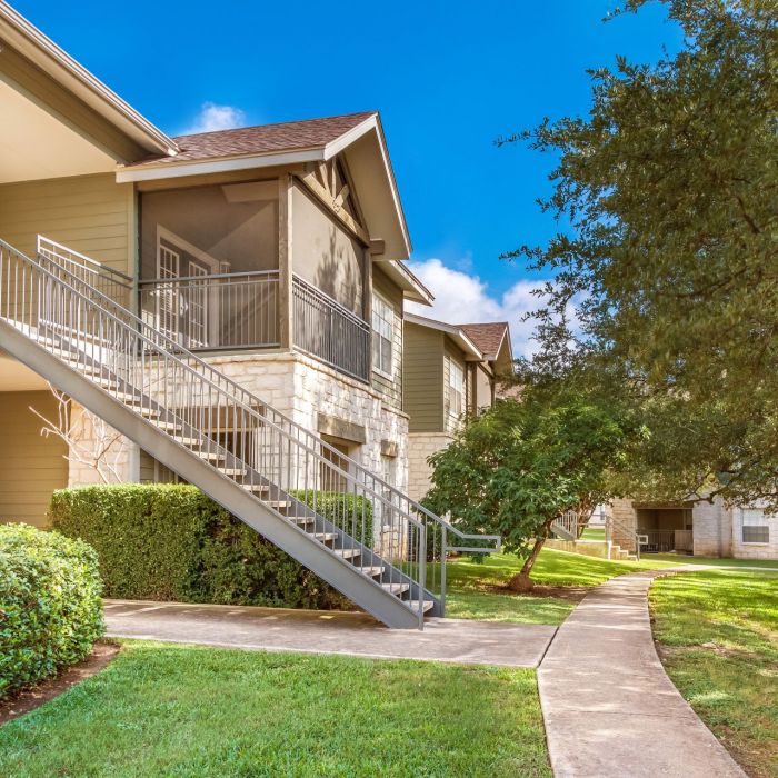 Elm Hollow Apartments boast exterior stairs leading up to charming units, surrounded by lush greenery and a scenic walkway, all basking in the warmth of a sunny day.