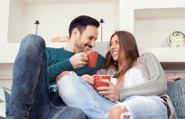 A couple at Elm Hollow Apartments sits on a cozy couch, smiling as they hold steaming mugs, with a clock ticking gently in the background.